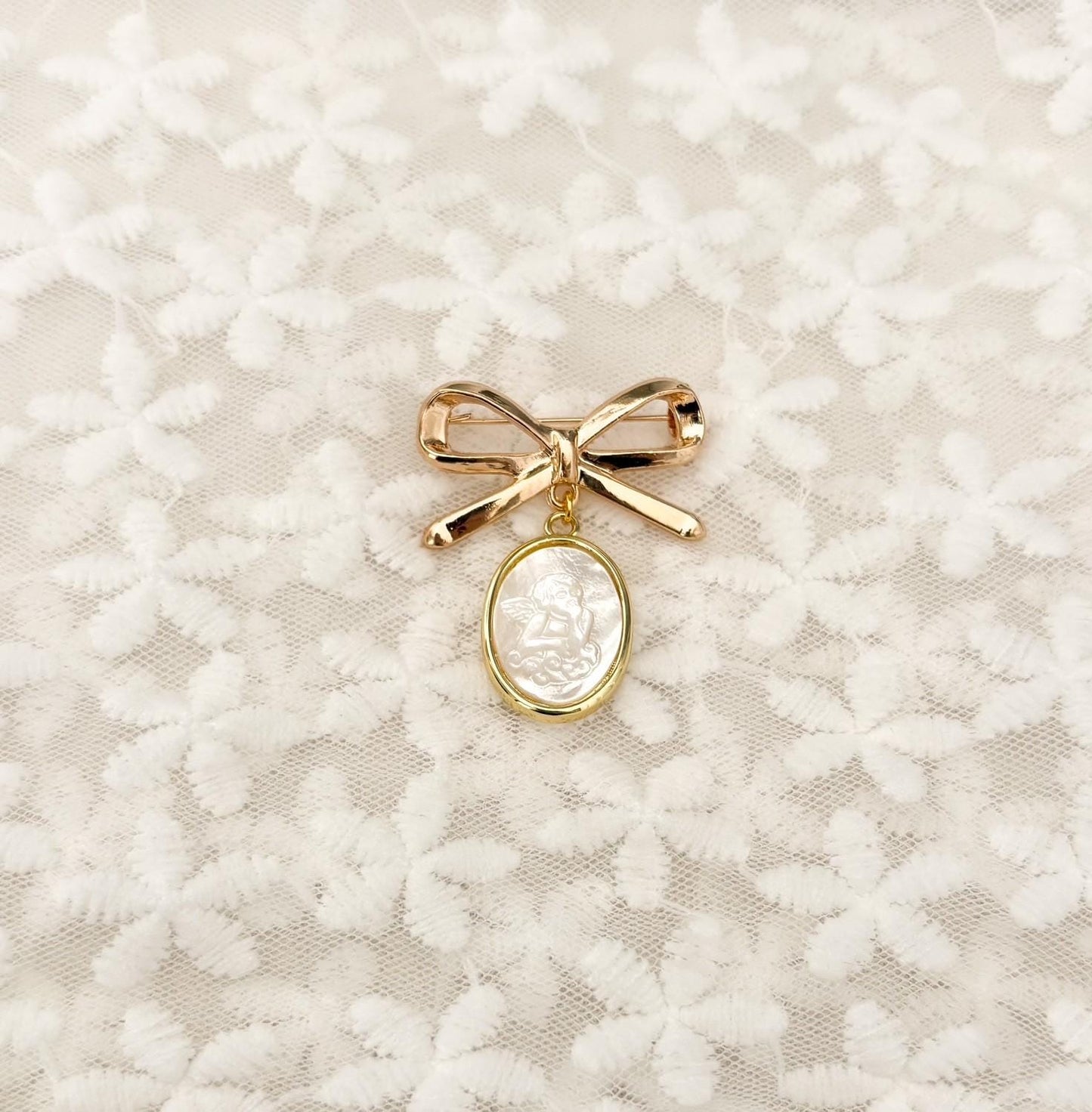 Delicate gold-tone bow baby brooch with dangling mother-of-pearl Guardian Angel charm – iridescent nacre angel medal set in gold rim, perfect Catholic baptism, christening, or blessing keepsake pin.