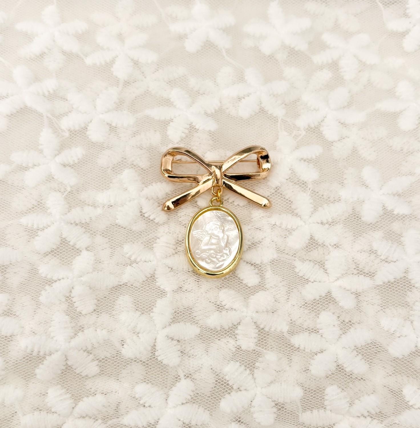 Delicate gold-tone bow baby brooch with dangling mother-of-pearl Guardian Angel charm – iridescent nacre angel medal set in gold rim, perfect Catholic baptism, christening, or blessing keepsake pin.
