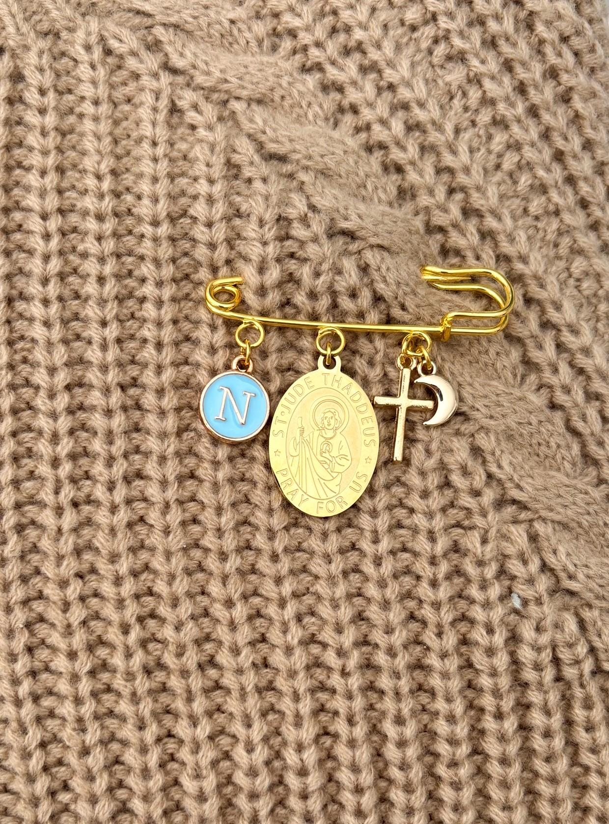 Elegant St Jude Baby Keepsake Pin with gold safety pin design, featuring a St Jude medallion, religious cross charm, personalised letter initial charm, presented in a luxurious Love & Grace pouch on a soft white background.
