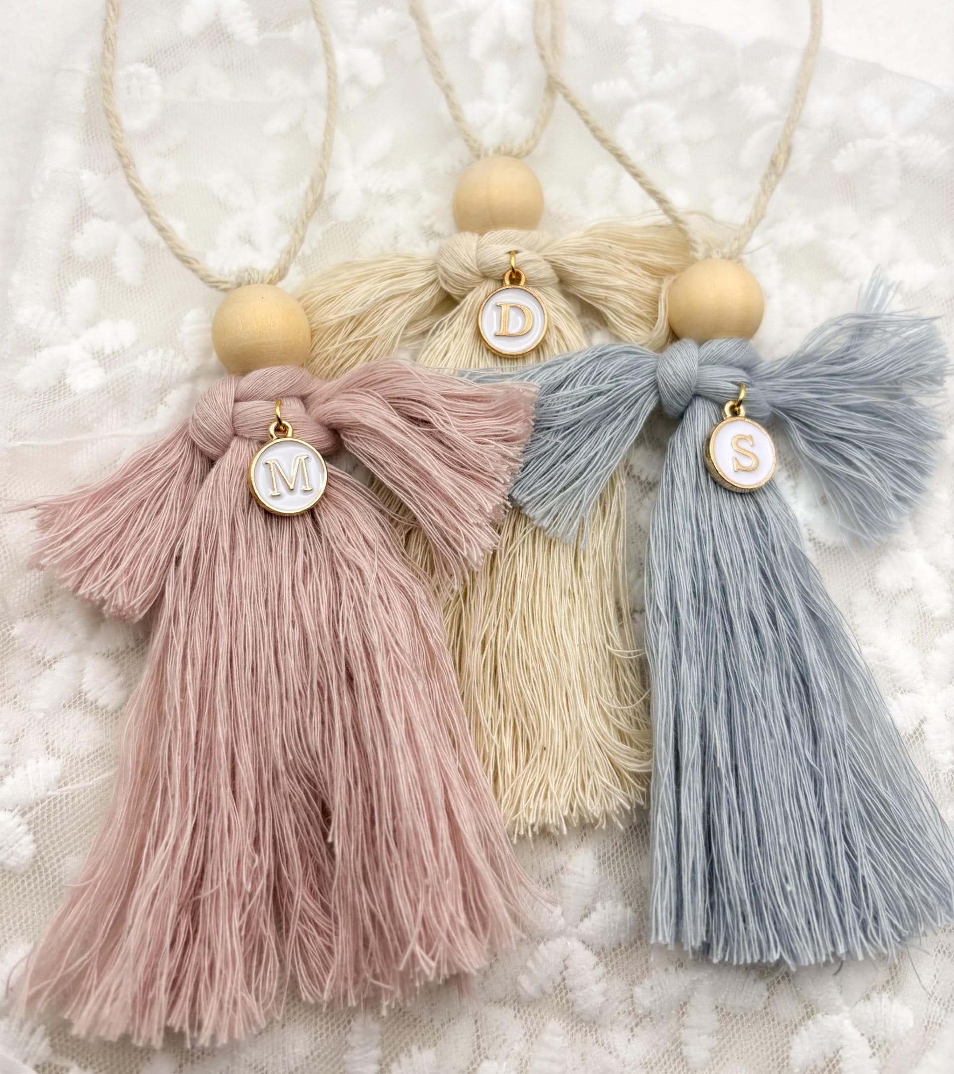Handmade Macrame Guardian Angel for Christening, Baptism, new baby Gift in Cream, Blue, or Pink. Tassel-style angels with wooden bead heads, featuring cream, blue, and pink yarn bodies. Each angel has a small circular charm with a personalised letter charm and is hung with a beige string. Set against a white lace background