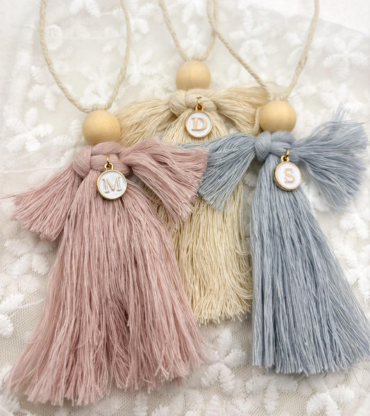 Handmade Macrame Guardian Angel for Christening, Baptism, new baby Gift in Cream, Blue, or Pink. Tassel-style angels with wooden bead heads, featuring cream, blue, and pink yarn bodies. Each angel has a small circular charm with a personalised letter charm and is hung with a beige string. Set against a white lace background